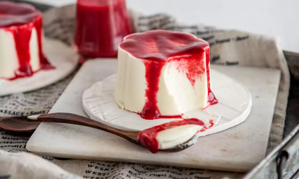 Picture - Šviesiai kreminės spalvos panna cotta cilindras, aplieta ryškiai raudonu tirštu padažu, padėta ant baltos lėkštės su mediniu šaukštu šalia