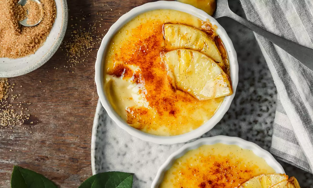 Picture - Dviejų baltų keramikinių indelių creme brulee su auksiniu karamelizuotu paviršiumi ir ananasų griežinėliais, padėtų ant gėlėtos lėkštės.